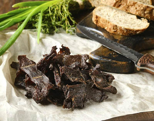 Beef jerky on parchment with herbs and bread on a rustic board.