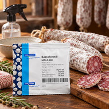 A package of CHR Hansen Bactoferm Mold-600 mold inhibitor for dried food, displayed on a rustic wooden cutting board with cured salami slices, hanging salami, a spray bottle, and rosemary.