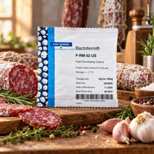 A single packet of Chr. Hansen Bactoferm F-RM-52 US fast fermenting culture stands on a wooden cutting board surrounded by sliced salami, garlic, rosemary, peppercorns, and other food props in a warm kitchen setting.