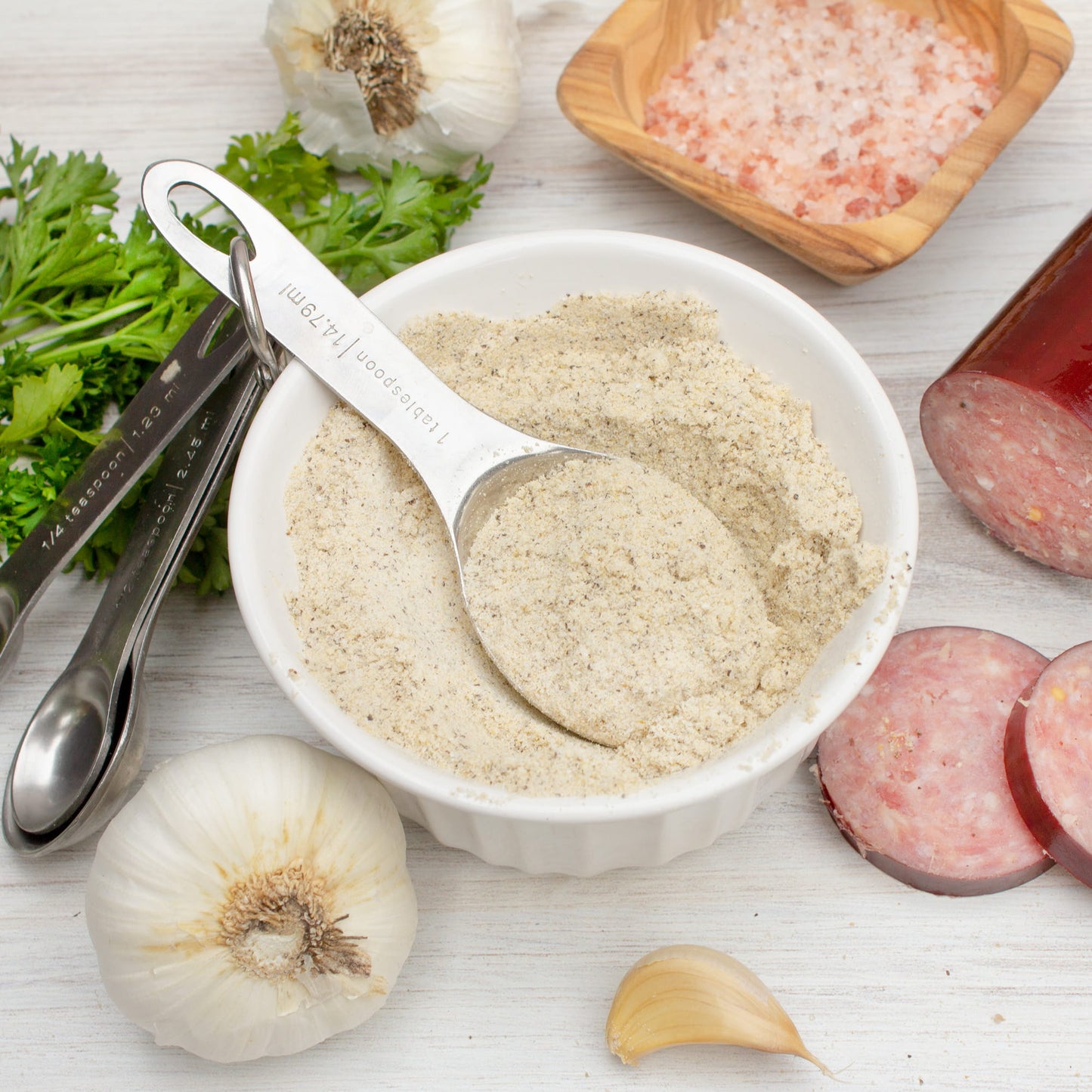Bowl of ground summer sausage seasoning with measuring spoons, garlic, and herbs on a wooden surface