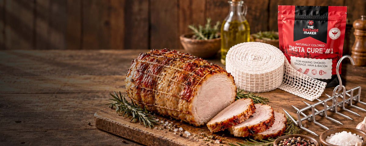 A sliced roasted meat loaf on a wooden cutting board with rosemary, next to Instant Cure #1 curing salt, a roll of meat netting, and a stainless steel bacon hanger on a rustic wooden table.
