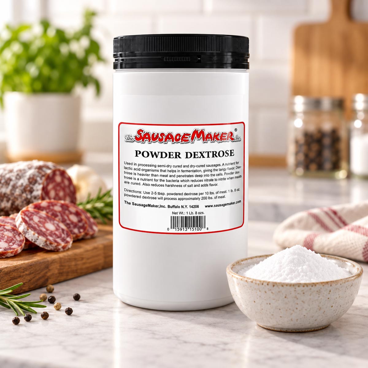 A white plastic tub of Sausage Maker Powder Dextrose with a black lid sits on a marble countertop, with sliced salami and a blurred kitchen background.